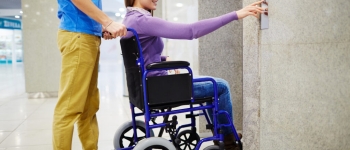 Assistant and handicapped young woman in wheelchair waiting for elevator, she pressing button by herself and smiling