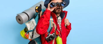 Studio shot of serious unshaven male backpacker keeps binoculars near eyes, wears hat and red jacket, explores new way, carries tourist backpack, stands against blue background, trekks somewhere